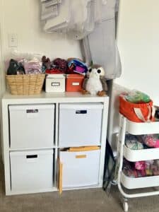An IKEA 4-shelf unit with an overflowing storage box in each unit. On top, there are baskets and piles of fabric.