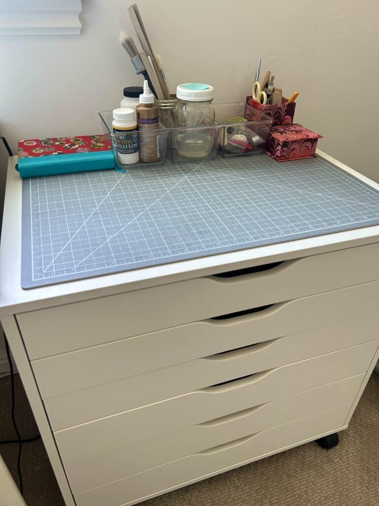 A small chest of flat drawers with a cutting mat on top of it. There are bins of glues and brushes in a line on top of the chest. 