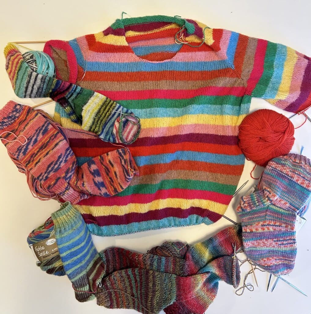 A 2/3 finished handknit sweater in rainbow stripes laid flat on a table. Five colorful handknit socks in progress surround it. 