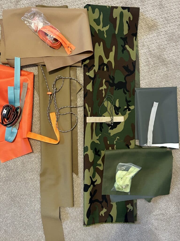 tan and camo outdoor fabrics piled on the floor, with bits of orange and tan webbing, paraford, and zippers on top.