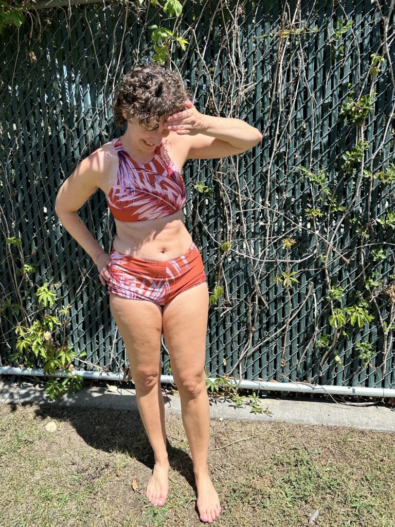 Portrait of a white middle aged woman in a high neck bikini top and bikini shorts. The swimsuit has a pattern of orange palm leaves. 
