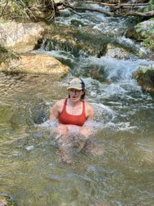 A white woman in a red sports bra and a baseball hat sits down in a mountain stream. Her mouth is open in a gasp.