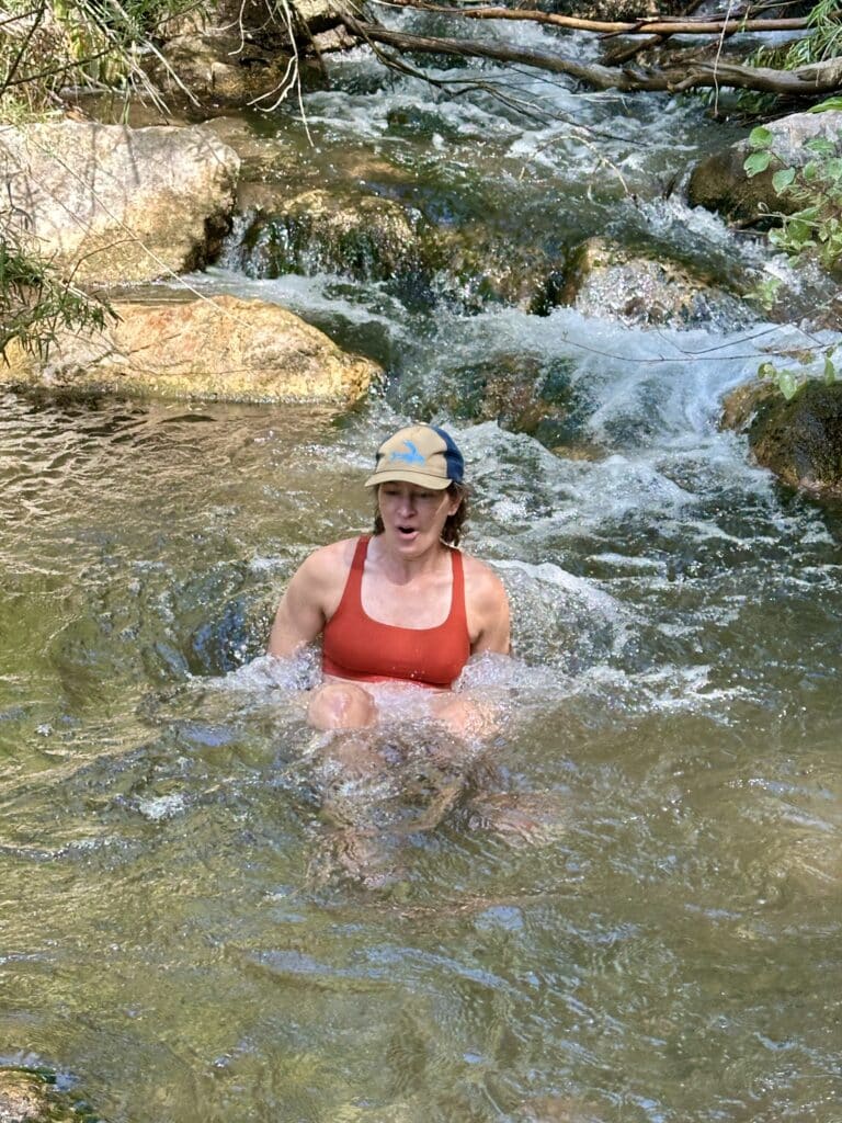 A white woman in a red sports bra and a baseball hat sits down in a mountain stream. Her mouth is open in a gasp. 