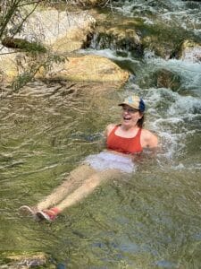 A white woman in a red sports bra and a baseball hat lies back in a mountain stream with her legs out. She is smiling but kind of wildly.