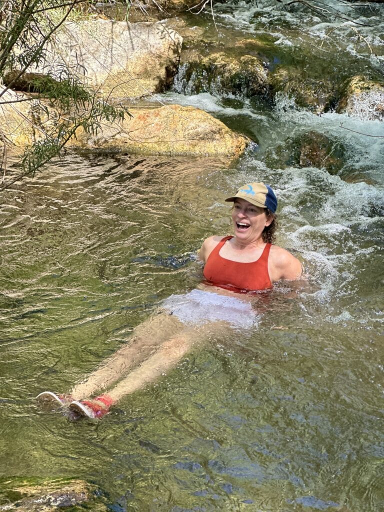 A white woman in a red sports bra and a baseball hat lies back in a mountain stream with her legs out. She is smiling but kind of wildly. 