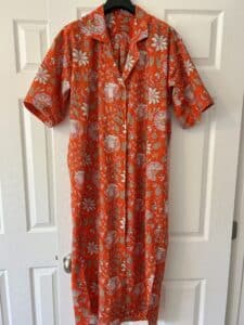 An orange and pink block print shirt dress with short sleeves displayed on a hanger over a white door.
