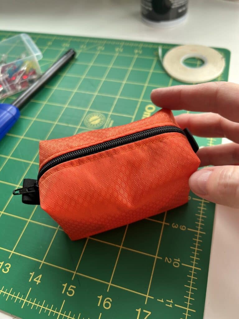 A hand holding a small orange pouch with a black zipper on a sewing table.
