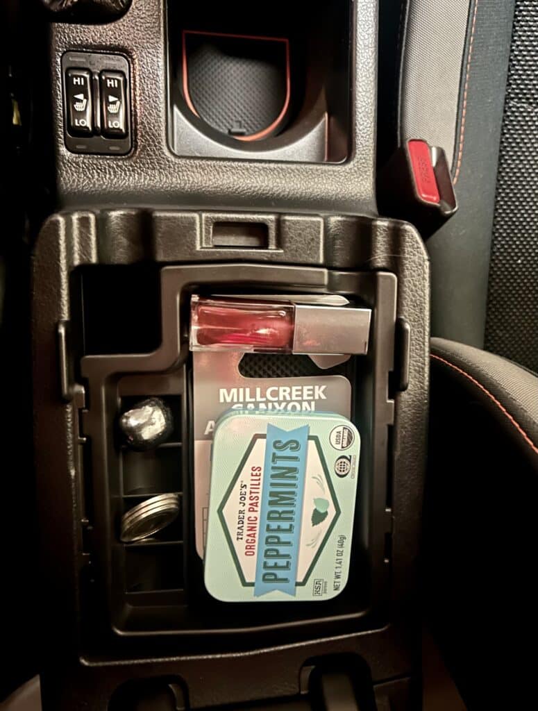 A top tray in a car's center console. There are mints and lip gloss and a canyon pass neatly arranged in it. 