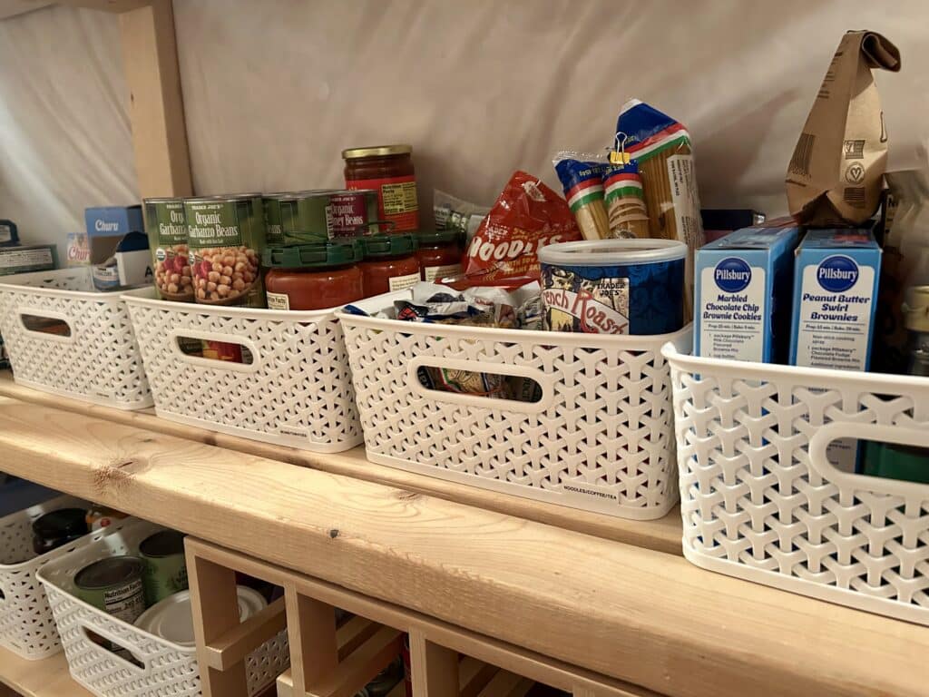 Labeled plastic bins full of cans and boxes sit on a wooden shelf in a row. 