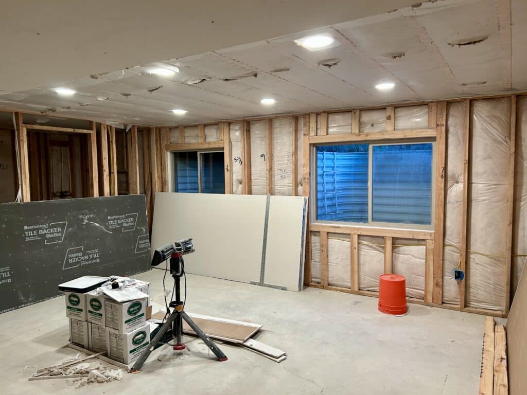 An unfinished basement with piles of drywall and cement board in it. Something looks off with the window. 
