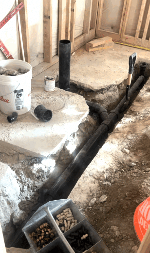 A framed basement with a deep trench with a sewer pipe in it. Tools and pickaxes are scattered around. 