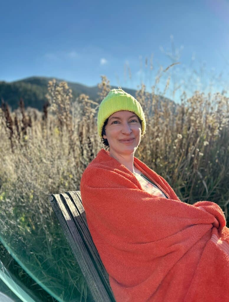A woman wearing a neon green wool hat, wrapped in a red towel, sitting at the side of the river. 