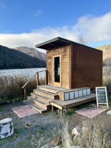 A wooden sauna building on the side of the river. It has a glass door and stairs going into it.