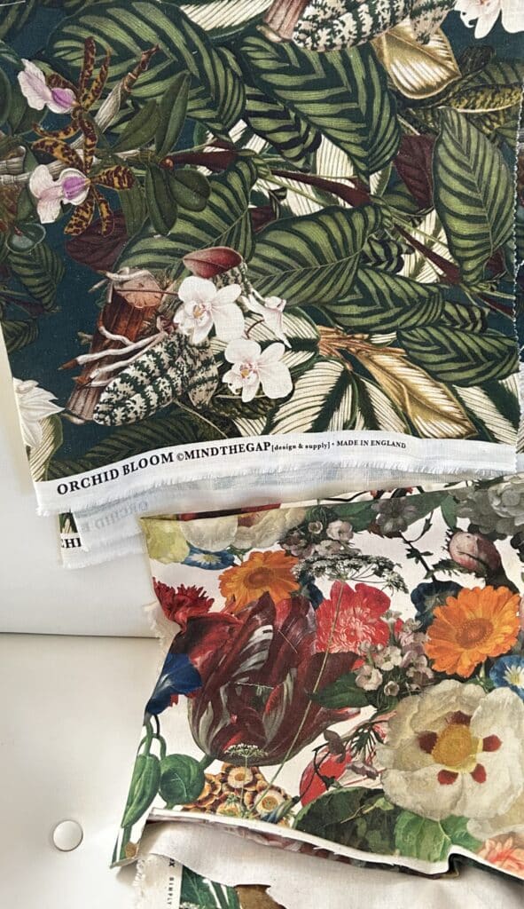 Two cuts of fabric draped over a white chair. One is a dark green print with orchids and the other is a rainbow Dutch floral