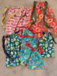 Nine fabric gift bags piled on a carpeted floor. They are bright prints of christmas ornaments and wreaths in red, pink, and turqupise.