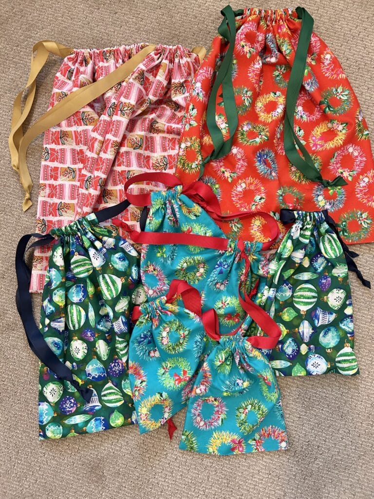 Nine fabric gift bags piled on a carpeted floor. They are bright prints of christmas ornaments and wreaths in red, pink, and turqupise.