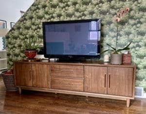 A mid-century style walnut-colored credenza with a TV and plants on it, in front of green tree-print wallpaper