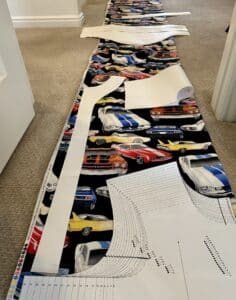 Pattern pieces on top of fabric stretching down a hallway. The fabric has a black background with primary-colored classic cars.