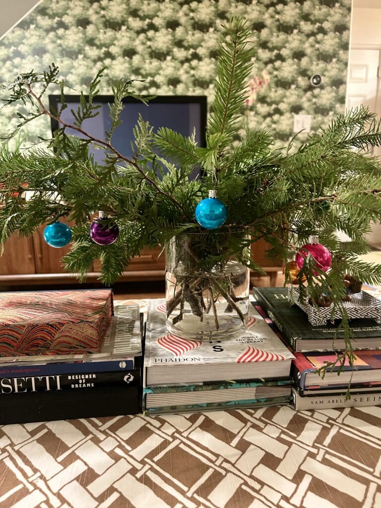A vase of evergreen branches hung with small colored balls sits on a coffee table covered in books.