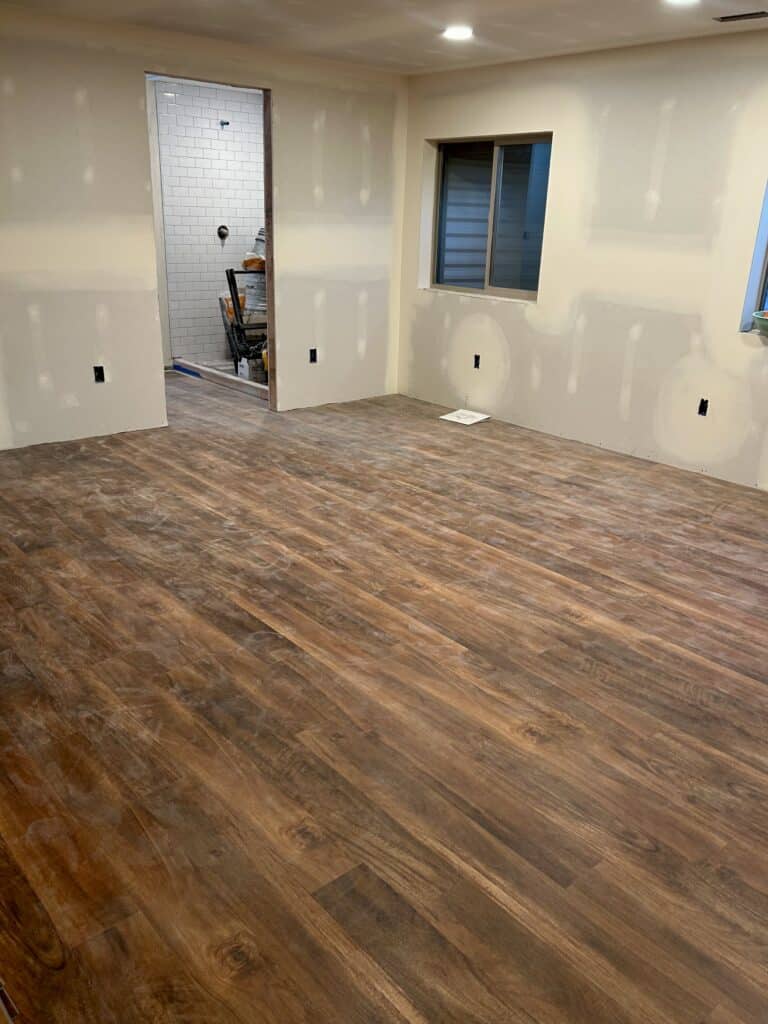 A dusty vinyl "wood" floor in a basement room. A bathroom with a tiled shower opens off of it.