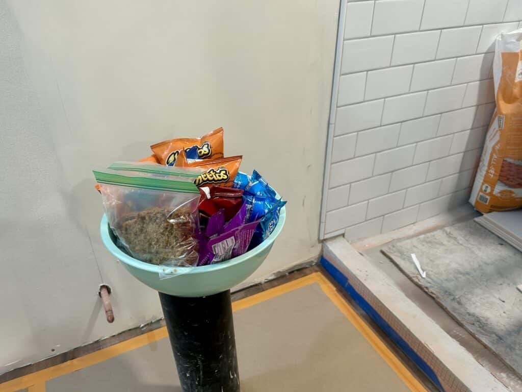 A bowl of chips and snacks sits on top of the stand pipe for the toilet in a bathroom.