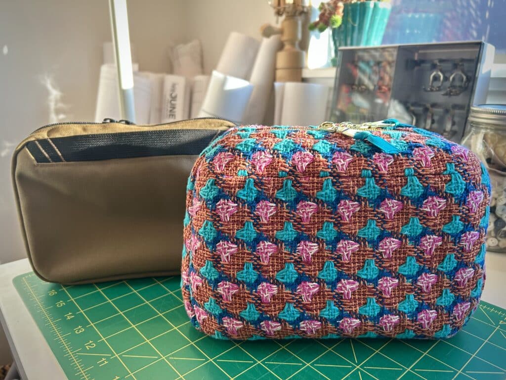 Two pouches on a sewing table. The one in front is made of orange, pink, and blue tweed. The one in back is made of tan technical fabric.