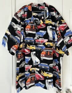 A mens shirt hanging in front of a white closet. It is black with red, yellow, and blue vintage muscle cars on it.