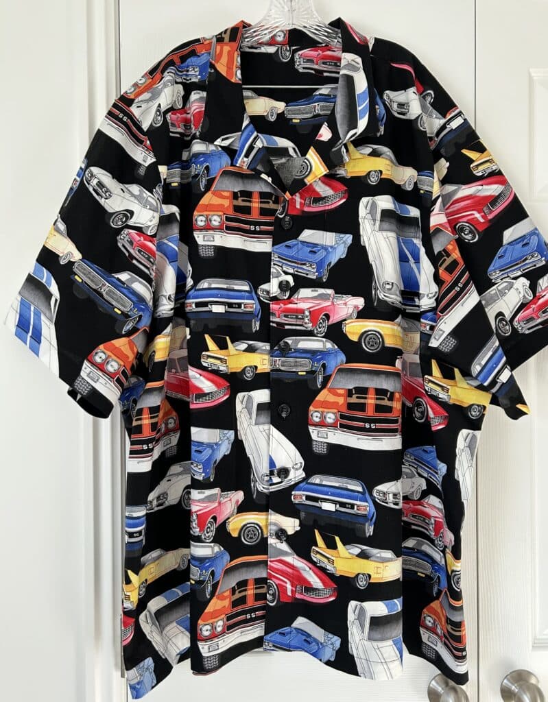 A mens shirt hanging in front of a white closet. It is black with red, yellow, and blue vintage muscle cars on it. 