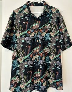 A men's shirt hanging in front of a white closet. It is black with turquoise and orange leis and volcanoes on it.