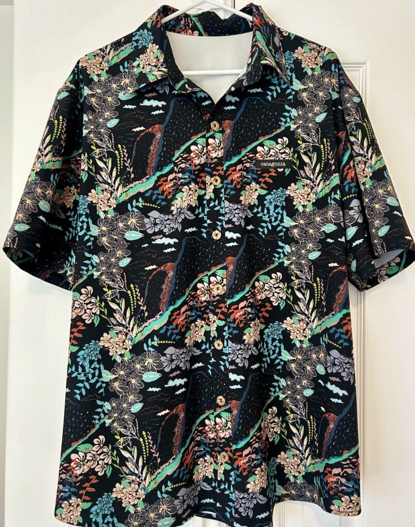 A men's shirt hanging in front of a white closet. It is black with turquoise and orange leis and volcanoes on it. 
