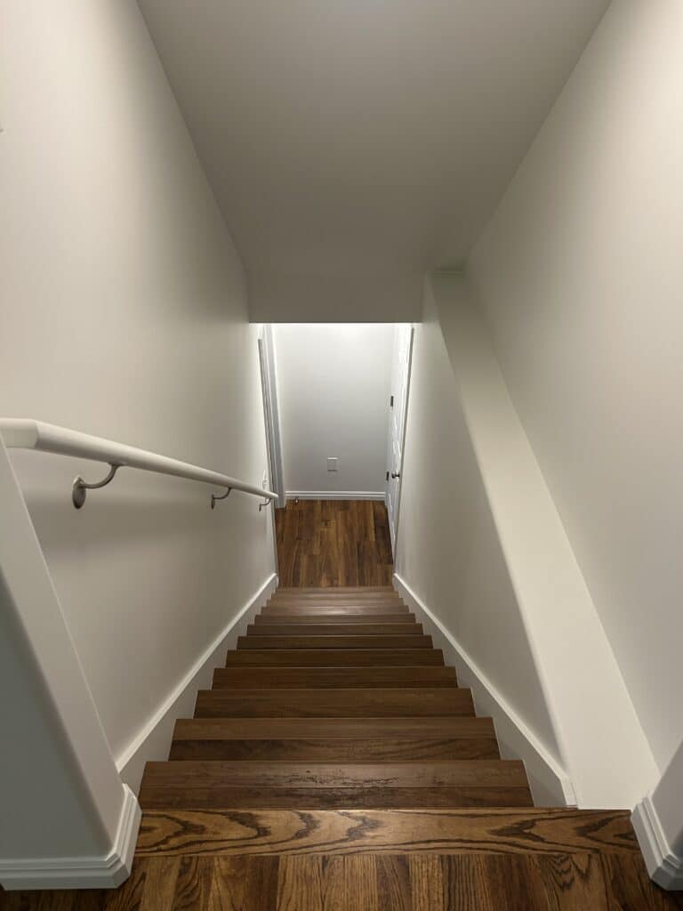 The same shot of the stairwell but with finished white walls, lighting, and wood-look stairs 