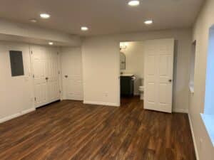 The same angle of the same basement, but now there are clean white walls, a bathroom, doors over the furnace, and wood-look flooring