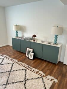 A long low cabinet with green-gray doors against a white wall. Two turquoise lamps are on it and a fluffy white rug is in the foreground.