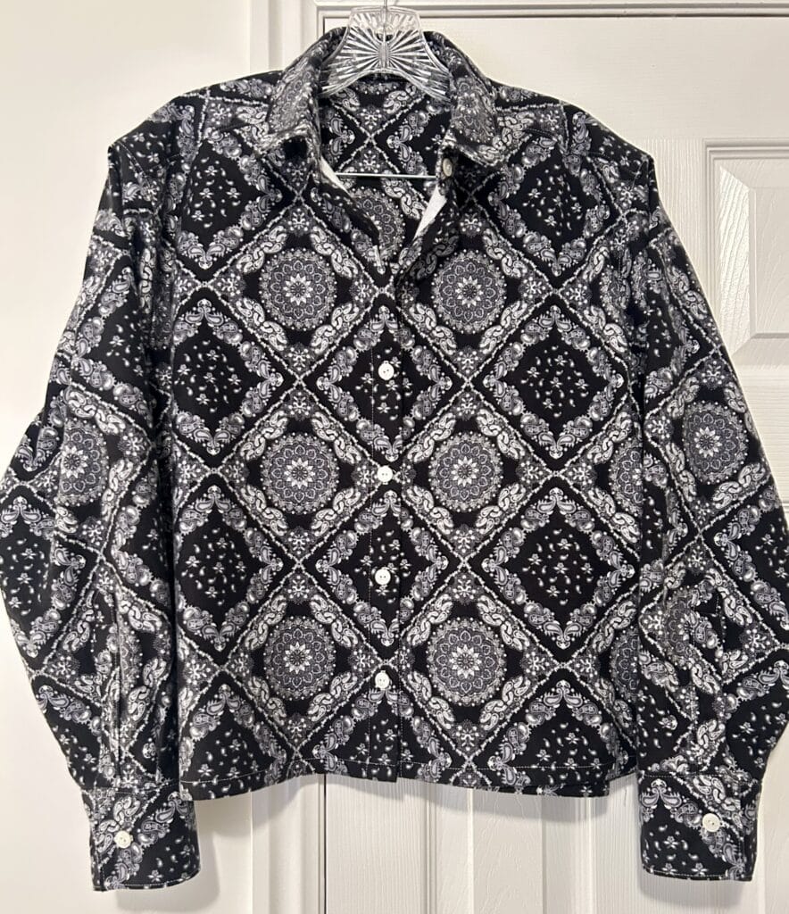 A cropped button up shirt in a white-on-black bandanna print hanging against a white closet door. 