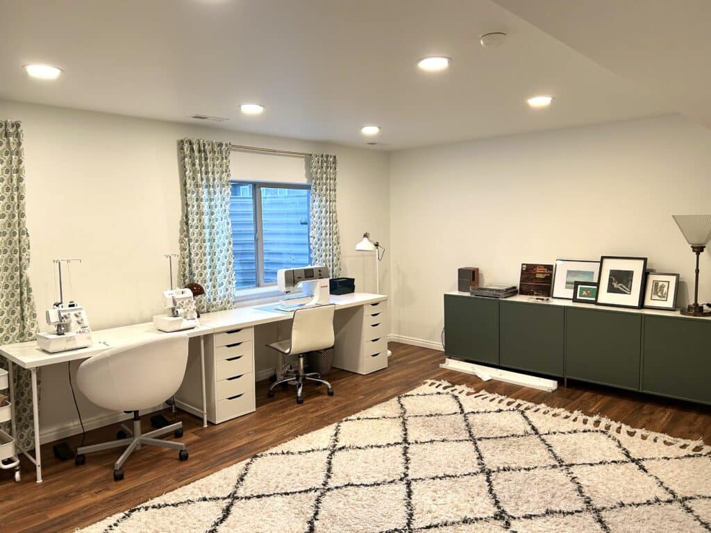 A large room with a long table with 3 sewing mahchines on it against one wall. A long green cabinet is against the other wall and there is a fluffy white rug. 
