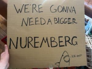 A cardboard sign with handwritten text and a small drawing of a shark. The text says, "WE'RE GONNA NEED A BIGGER NUREMBERG" and the shark is saying, "ICE OUT"
