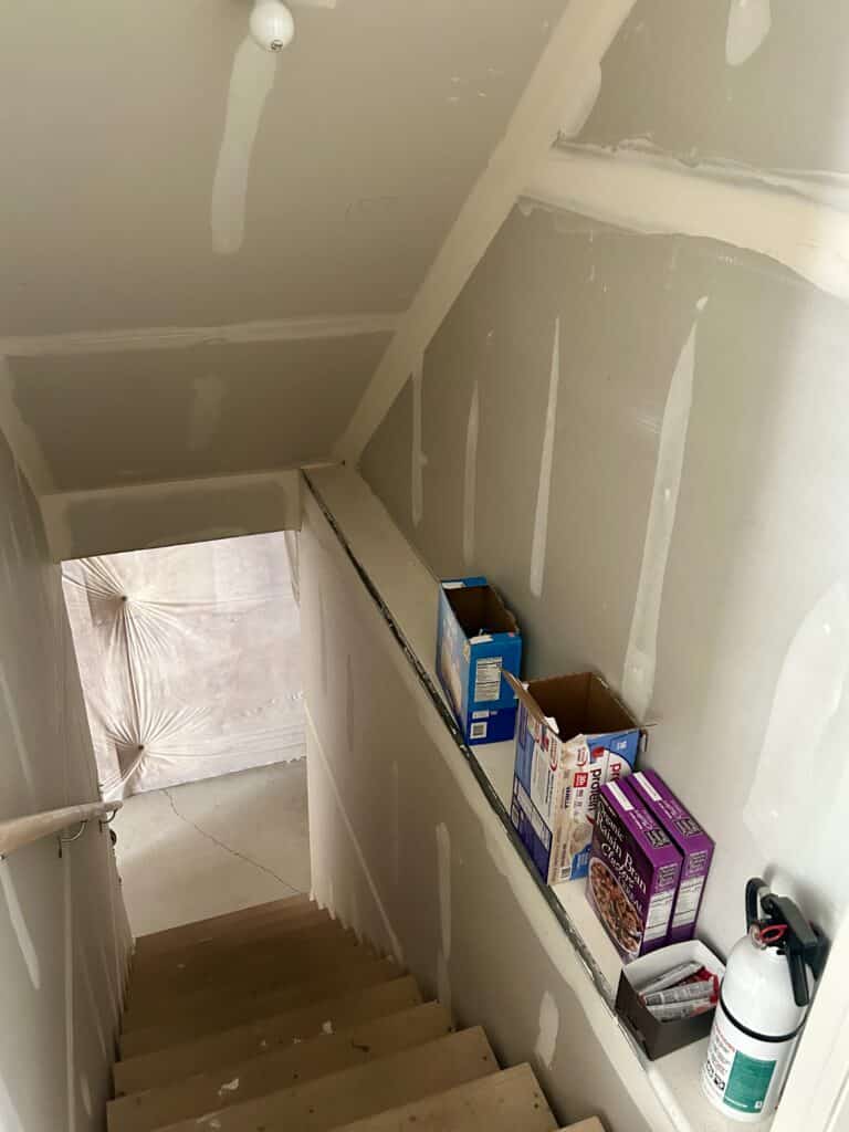 An unfinished stairwell leading into the basement. Various pantry items are stacked on the stair ledge. 