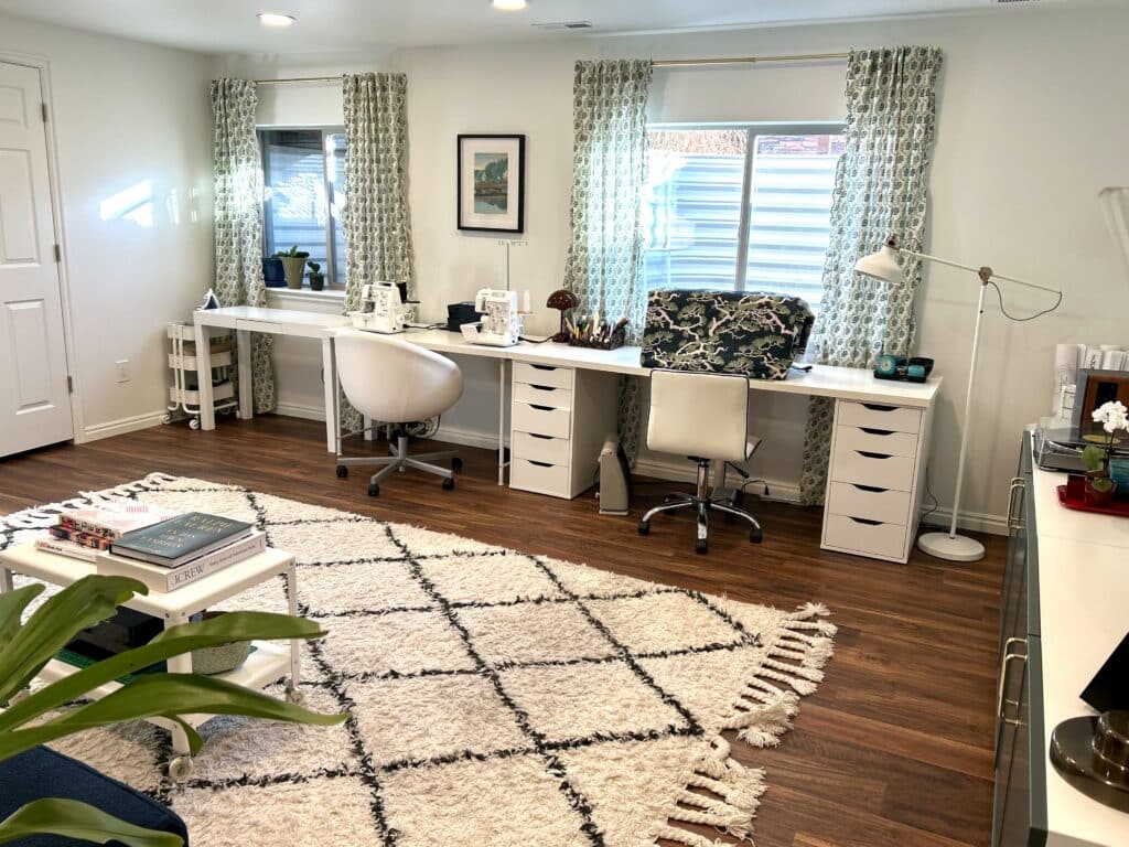 Three long white desks against a wall with two curtained windows in it. The desks have three sewing machines on them. 