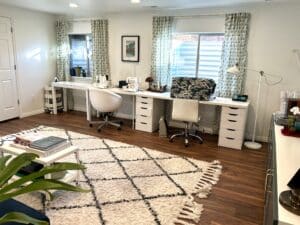 Three long white desks against a wall with two curtained windows in it. The desks have three sewing machines on them.