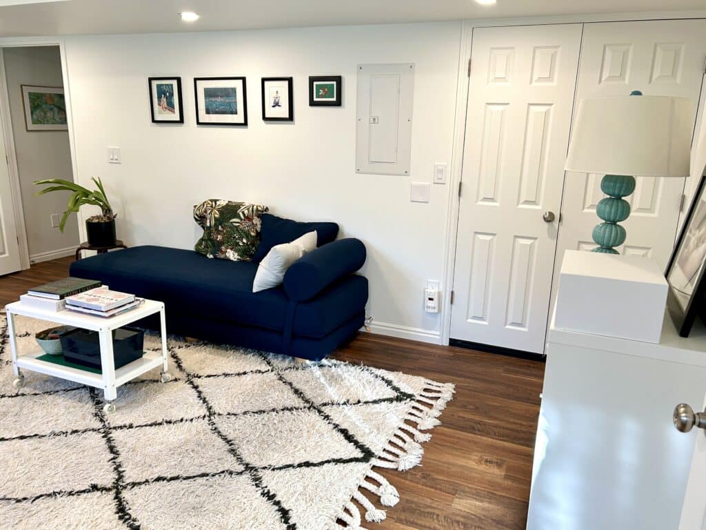 A navy blue chaise lounge with a white coffee table on wheels in front of it. There is a fluffy white rug on the floor. 
