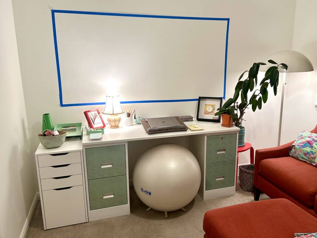 A desk and filing cabinet against a white wall with an orange coach along the other wall. Above the desk a 6x3 foot rectangle is taped out in blue tape.