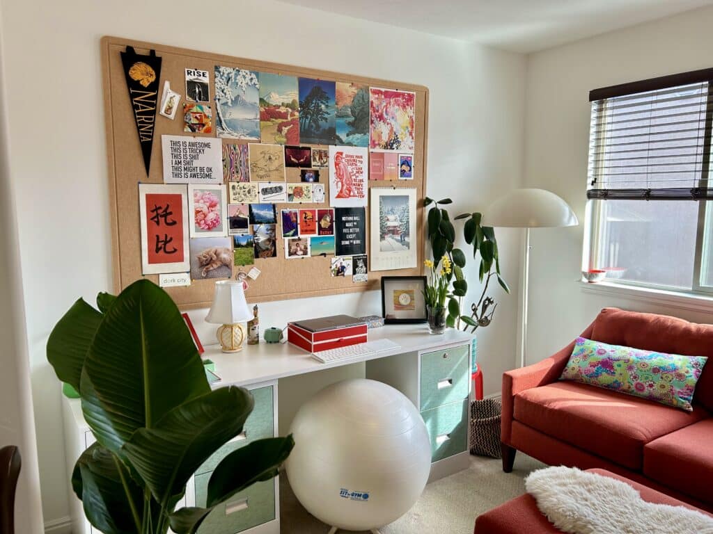 A desk with a large pinboard over it. The board is covered in bright posters and postcards.