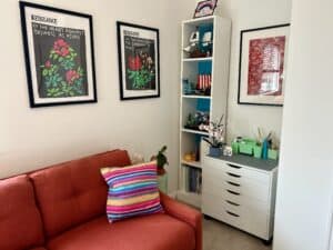 A corner of a white room with posters on the walls and a tall shelf displaying LEGO kits. An orange couch is in the foreground with a bright striped knit throw pillow on it.