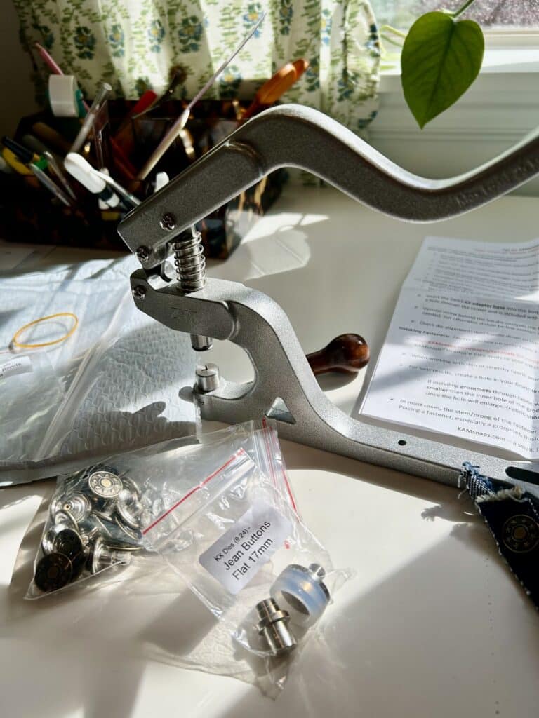 A metal table press for installing hardware on a work surface, surrounded by dies and bags of rivets and buttons.