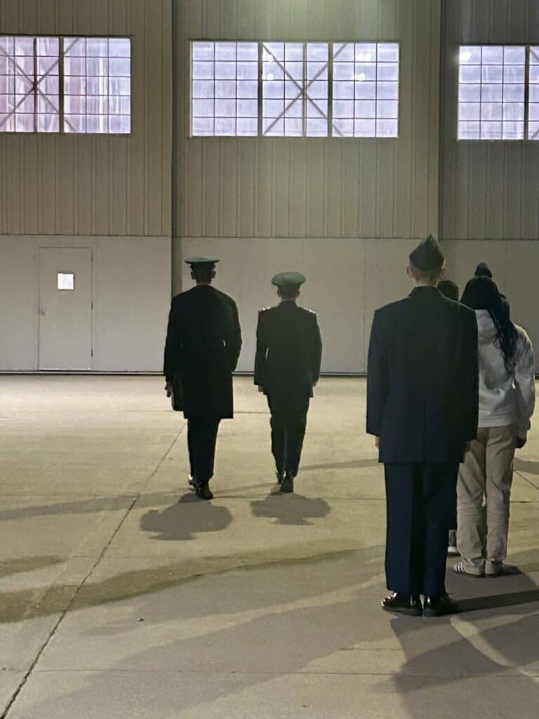 Two cadets march towards a hanger with lighted windows. The cadet on the left is taller. 