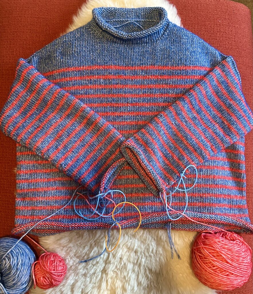 An almost-finished washed blue sweater with red stripes laid out on on orange ottoman.