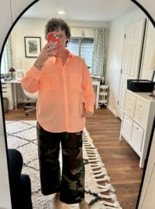 A woman takes a mirror selfie. She's wearing a neon apricot striped shirt, camo pants, and clogs.