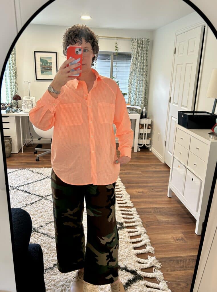 A woman takes a mirror selfie. She's wearing a neon apricot striped shirt, camo pants, and clogs. 