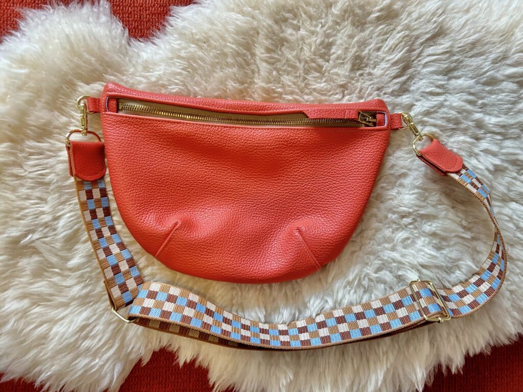 A homemade poppy red leather pouch with a tan checkered strap, laying on a fluffy white sheepskin