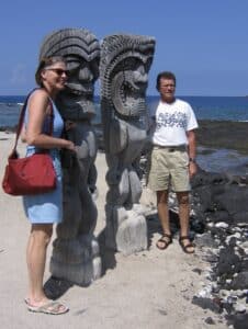 A woman and man in their 50s stand on a beach next to two carved Hawaiian figures